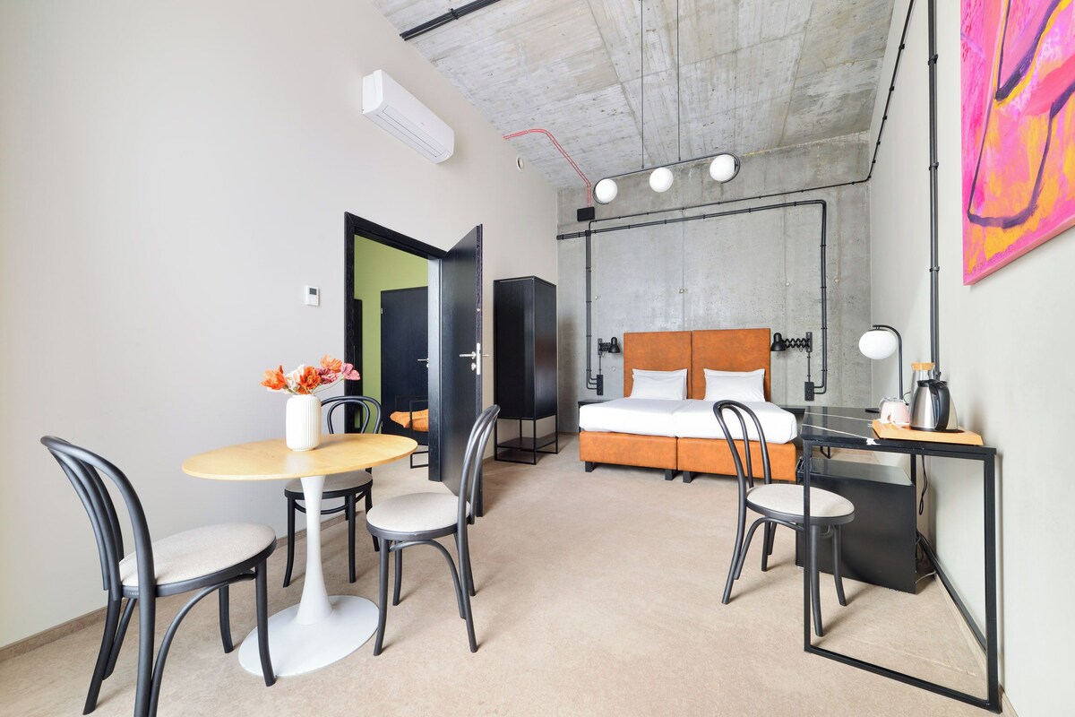 A modern hotel room with a bed, a desk, a chair, a table, and a small kitchenette.