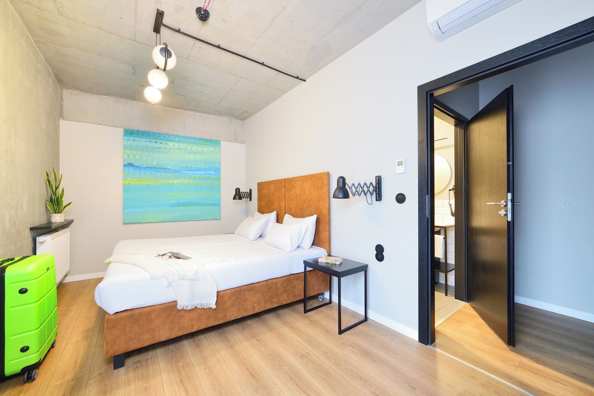 A modern bedroom with a wooden bed, a small bedside table, a bright green suitcase, and a painting on the wall.