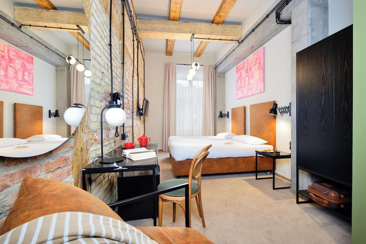 A modern hotel room with exposed brick walls, a loft bed, a desk with a chair, and a large wardrobe.
