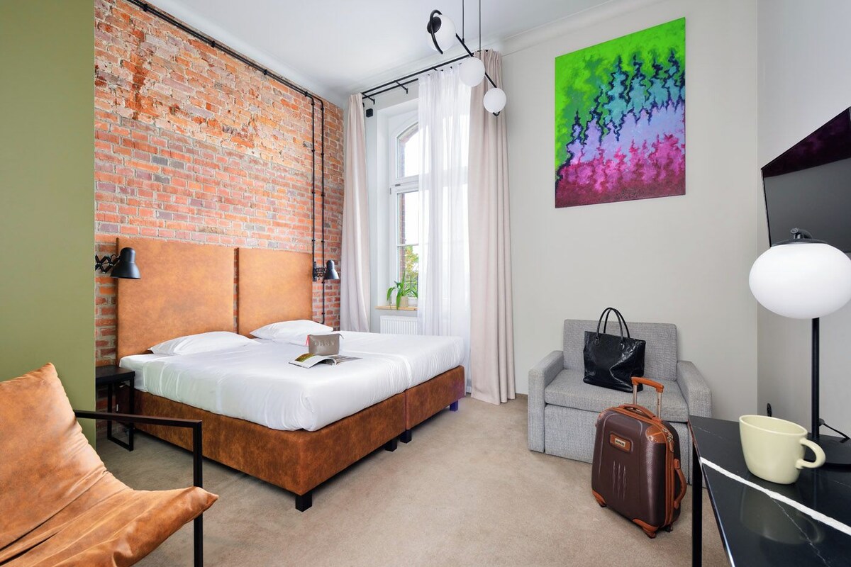 A hotel room with a brick wall, a bed, a grey sofa, a desk with a lamp, a suitcase, and a colorful painting on the wall.