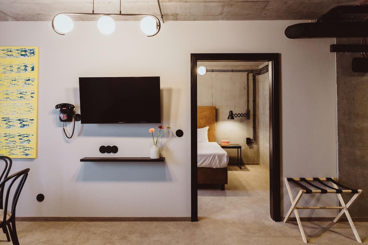 Una habitación de hotel moderna con una televisión de pantalla plana montada en la pared, un escritorio pequeño con un teléfono, una mesita de noche con una lámpara y un tendedero de madera.