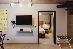 Una habitación de hotel moderna con una televisión de pantalla plana montada en la pared, un escritorio pequeño con un teléfono, una mesita de noche con una lámpara y un tendedero de madera.