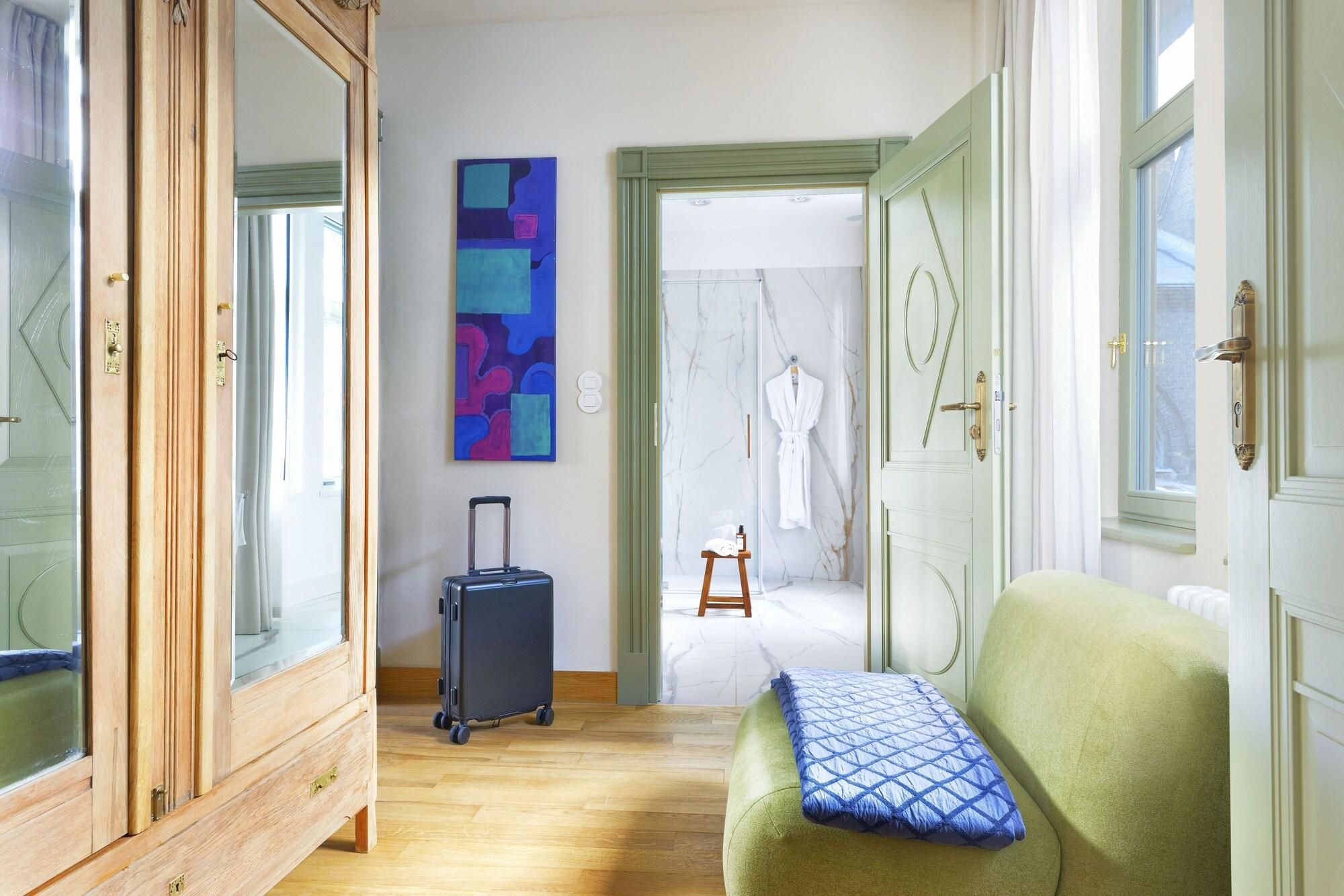 Un pasillo de hotel con un armario de madera, un cuadro abstracto azul, una maleta negra, un sofá verde con un cojín azul, y un baño visible a través de una puerta abierta.