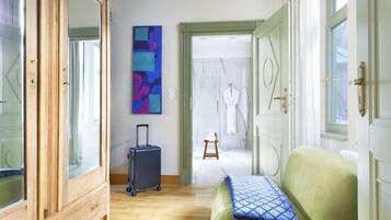 Un pasillo de hotel con un armario de madera, un cuadro abstracto azul, una maleta negra, un sofá verde con un cojín azul, y un baño visible a través de una puerta abierta.