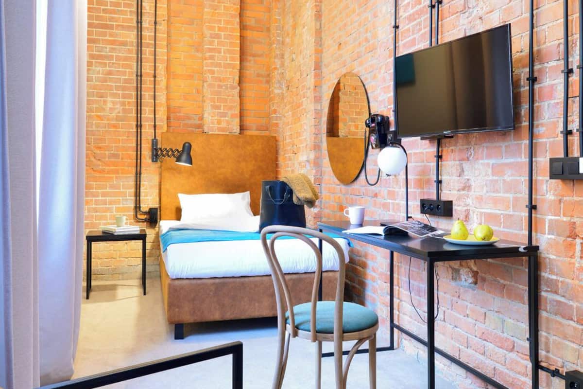 A modern hotel room with a brick wall, a bed, a desk, a chair, a TV, and a window with curtains.