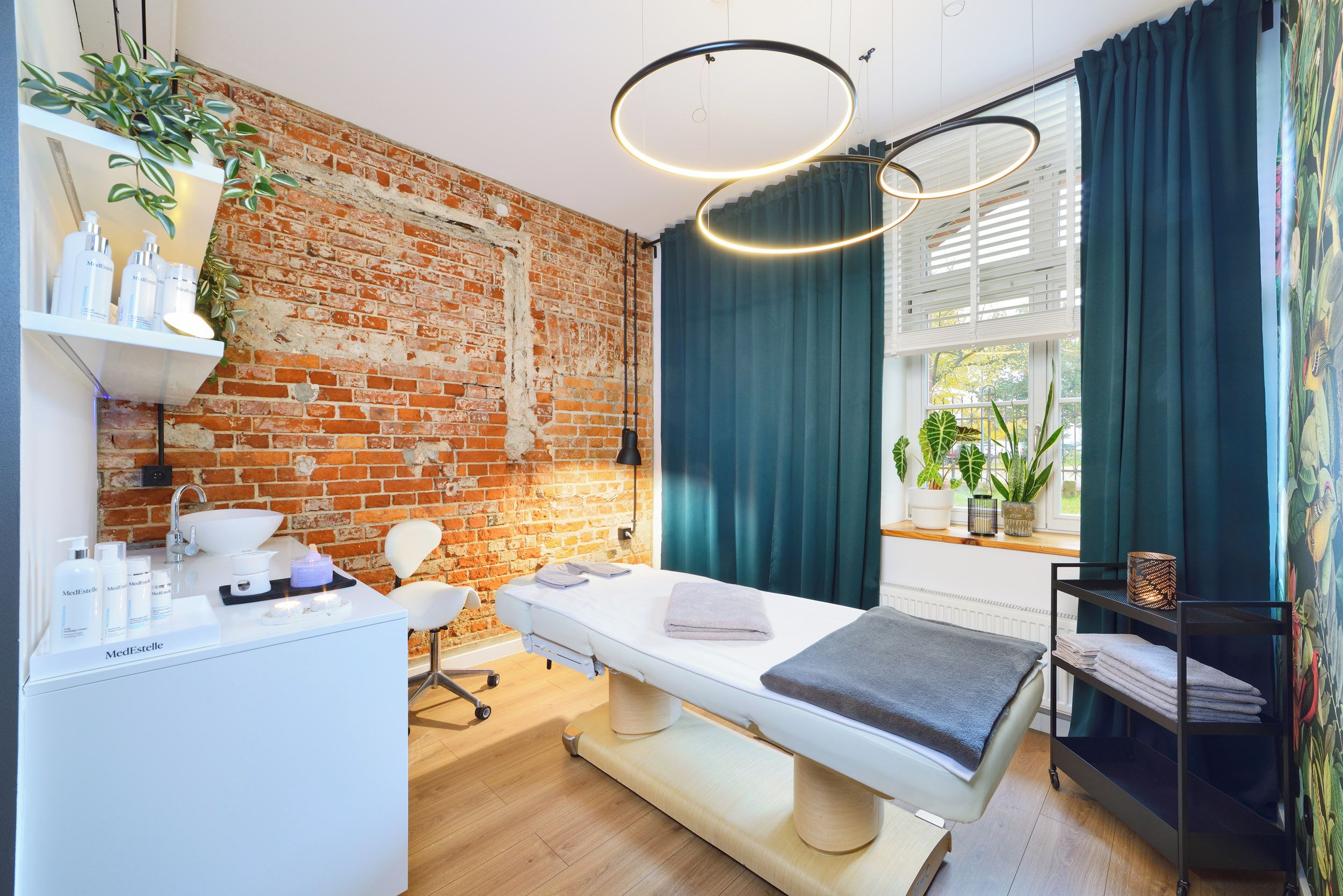 Ein moderner Spa-Raum mit Backsteinwand, einem weißen Empfangstresen, einer Massageliege und einem Fenster mit Vorhängen.