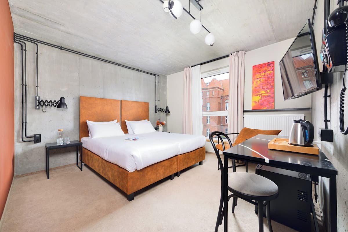 A hotel room with a bed, a desk, a chair, a TV, and a window with a view of buildings.