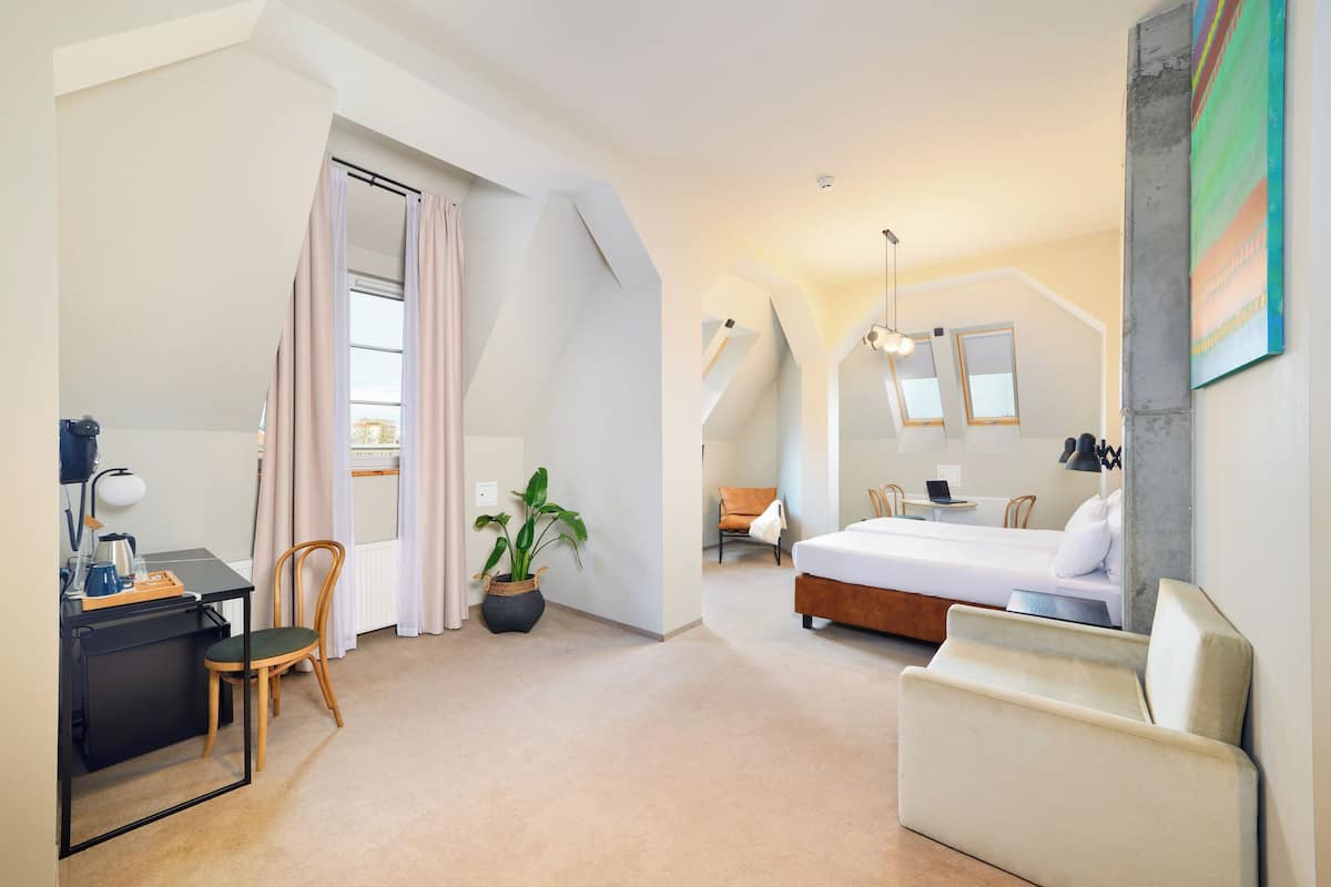 A modern hotel room with a bed, a desk, a chair, a potted plant, and a sofa.