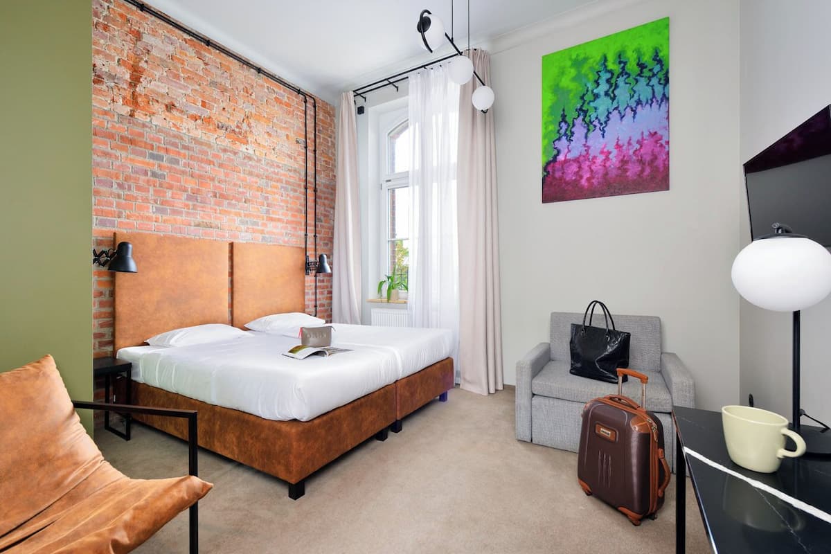 A hotel room with a brick wall, a bed, a grey sofa, a desk with a lamp, a suitcase, and a colorful painting on the wall.