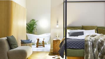A modern bedroom with a wooden floor, a green sofa, a white bathtub, and a bed with multiple pillows.