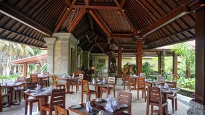 An outdoor dining area with wooden tables and chairs, stone columns, and a view of greenery. - Sakti Garden Resort & Spa (Ubud)