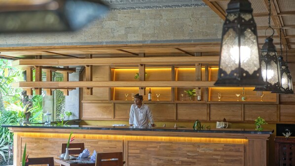 Un espace bar en bois, avec des étagères où sont exposés des verres et des bouteilles, un comptoir derrière lequel se tient une personne, et un luminaire suspendu.
