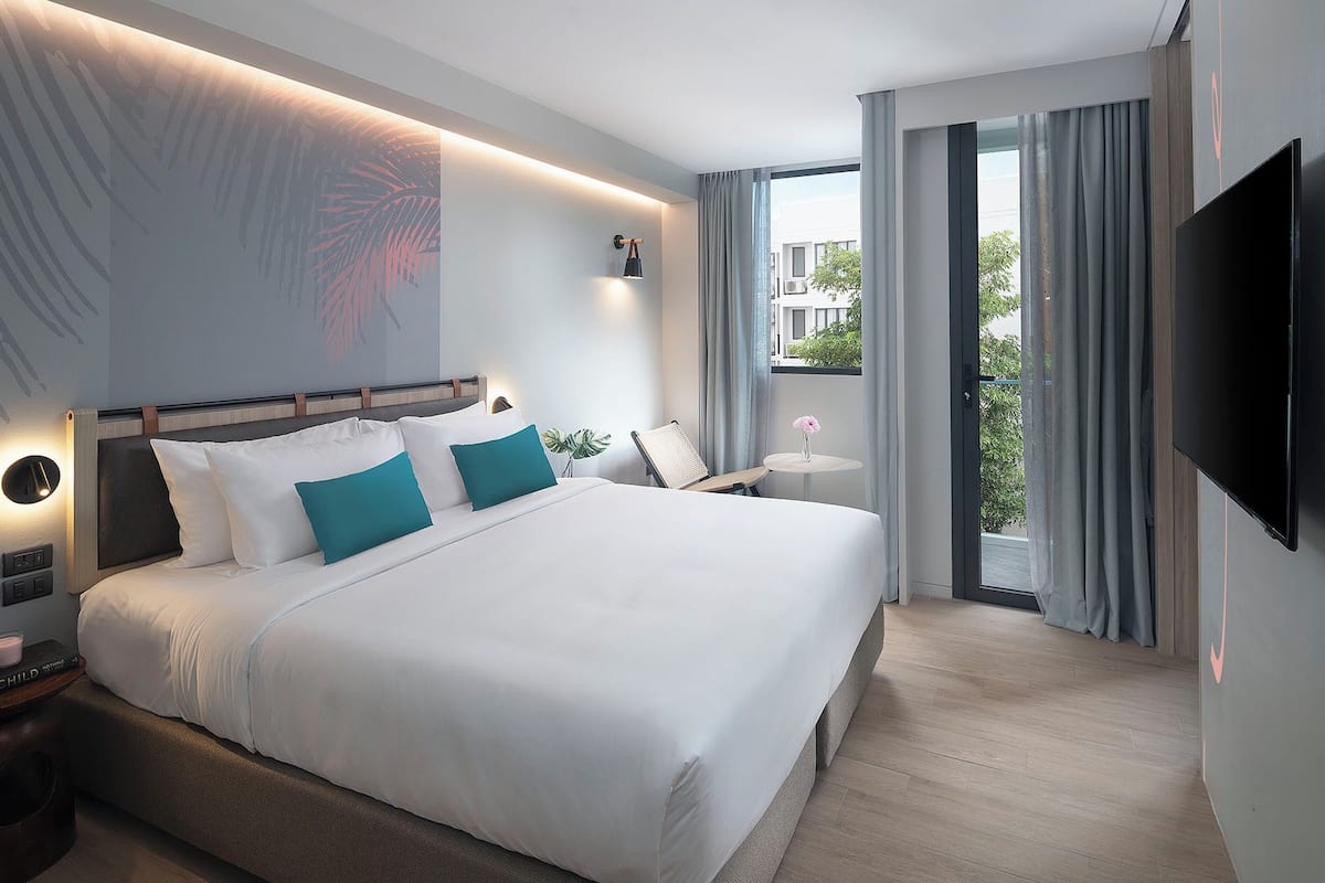 A modern hotel room with a large bed, a bedside table, a chair, a small table with a vase, and a TV.