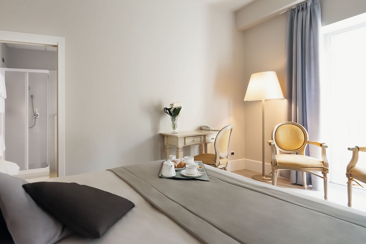 Hotellihuone, jossa on sänky, yöpöydällä lamput, työpöytä tuolilla, peilillä varustettu meikkipöytä ja ikkuna, jossa on verhot.