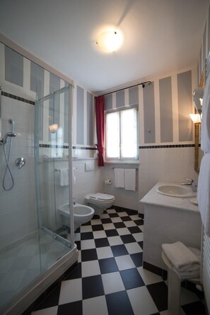 A bathroom with a glass shower enclosure, a toilet, a sink, and a window with curtains. - Hotel Cannero (Cannero Riviera)