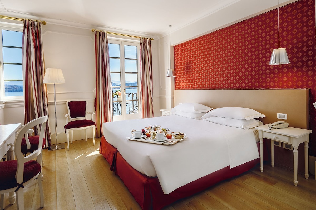 A hotel room with a bed, a desk, a chair, a bedside table with a lamp, and a view of the outdoors.