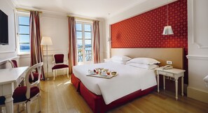 A hotel room with a bed, a desk, a chair, a bedside table with a lamp, and a view of the outdoors. - Hotel Cannero (Cannero Riviera)