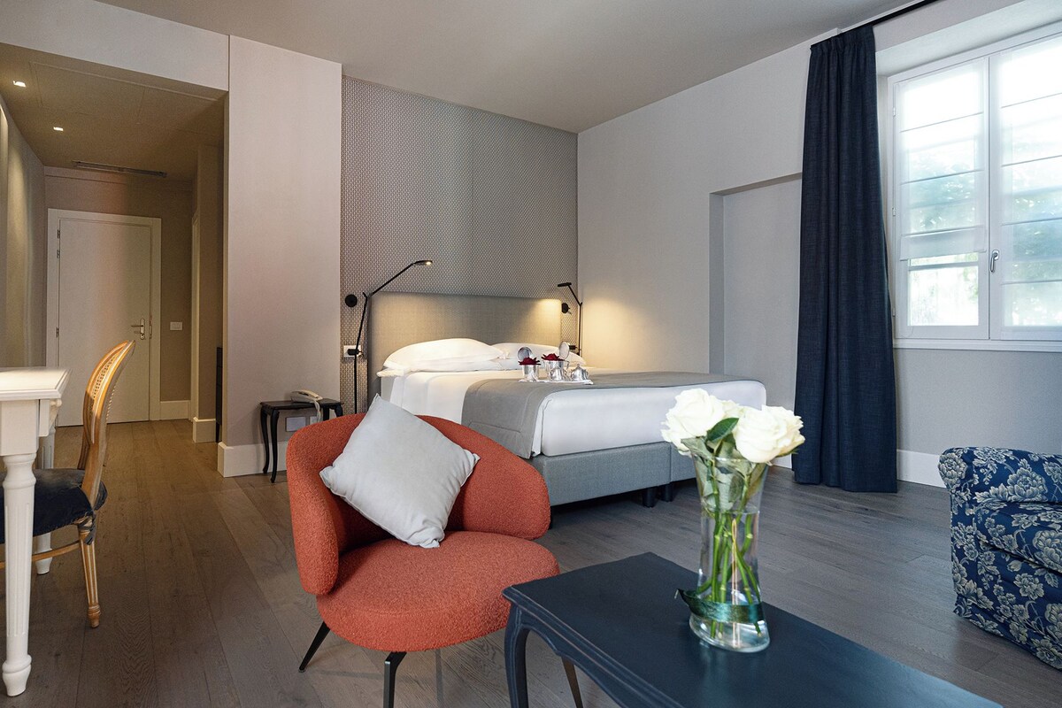 A hotel room with a bed, a desk with a lamp, a chair, a sofa, and a vase with flowers.