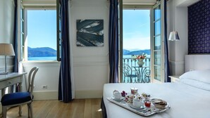 A hotel room with a bed, a desk, a chair, a balcony with a view of the sea, and a tray with breakfast items. - Hotel Cannero (Cannero Riviera)