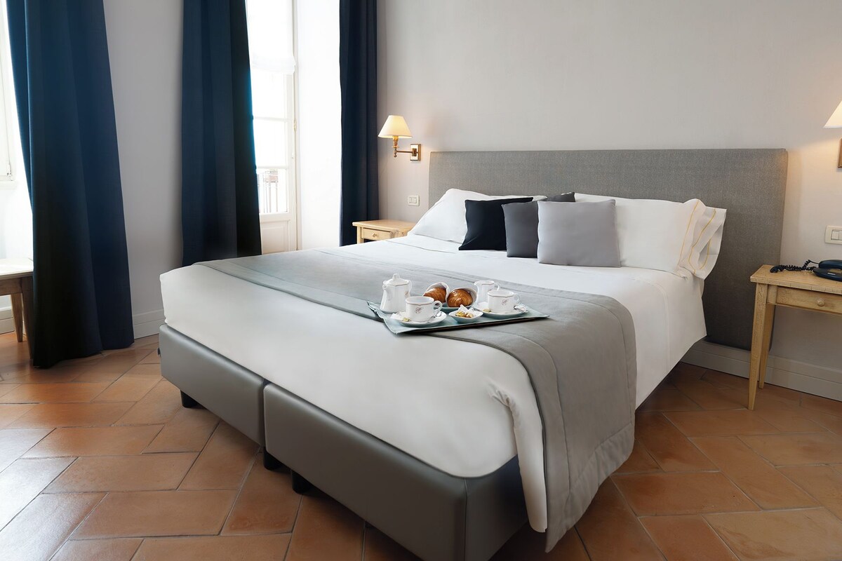 A neatly made bed with a tray of breakfast items, two bedside tables with lamps, and a window with curtains.