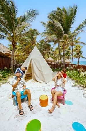Escena de playa con una carpa, palmeras y gente sentada en sillas de playa.
