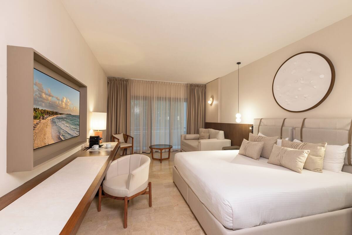 Habitación de hotel con una cama grande, un escritorio con una silla, una mesita pequeña y un televisor mostrando una escena de playa.