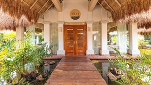 A wooden entrance with a thatched roof, a sign reading "The Nest", and a pathway leading to a water feature. - Family Selection At Grand Palladium Kantenah (Kantenah)
