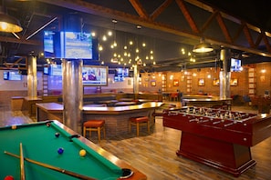 Eine Bar mit einer Billardtisch, einem Tischfußballtisch und mehreren Fernsehern, die Sportübertragungen zeigen.