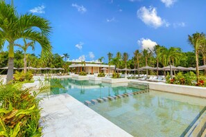 Zona de piscina con sillones de descanso y sombrillas, rodeada de palmeras y un edificio con fachada blanca.