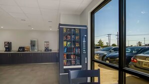 A modern indoor setting with a vending machine, a counter with a coffee machine, and a view of parked cars through large windows.
