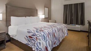 Ein Hotelzimmer mit einem großen Bett, einem Nachttisch, einer Lampe, einem Fenster mit Vorhängen und einer Wand-Klimaanlage.