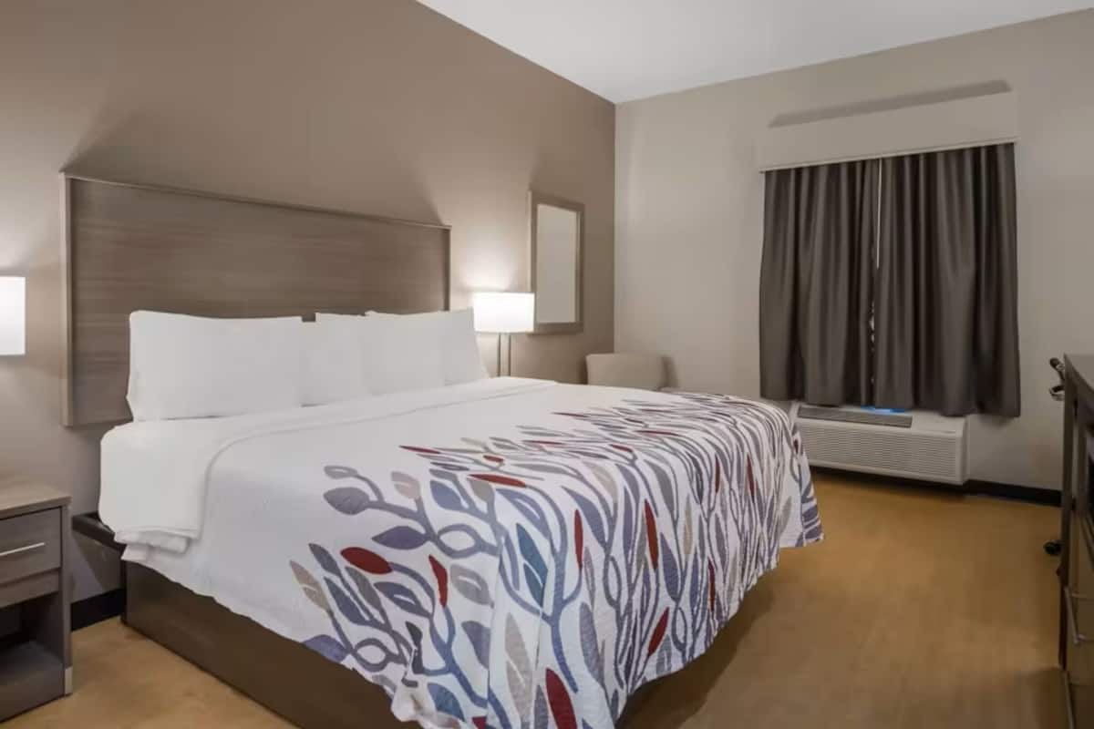 Quarto de hotel com uma cama grande, uma mesa de cabeceira, uma luminária, uma janela com cortinas e um ar condicionado fixado na parede.