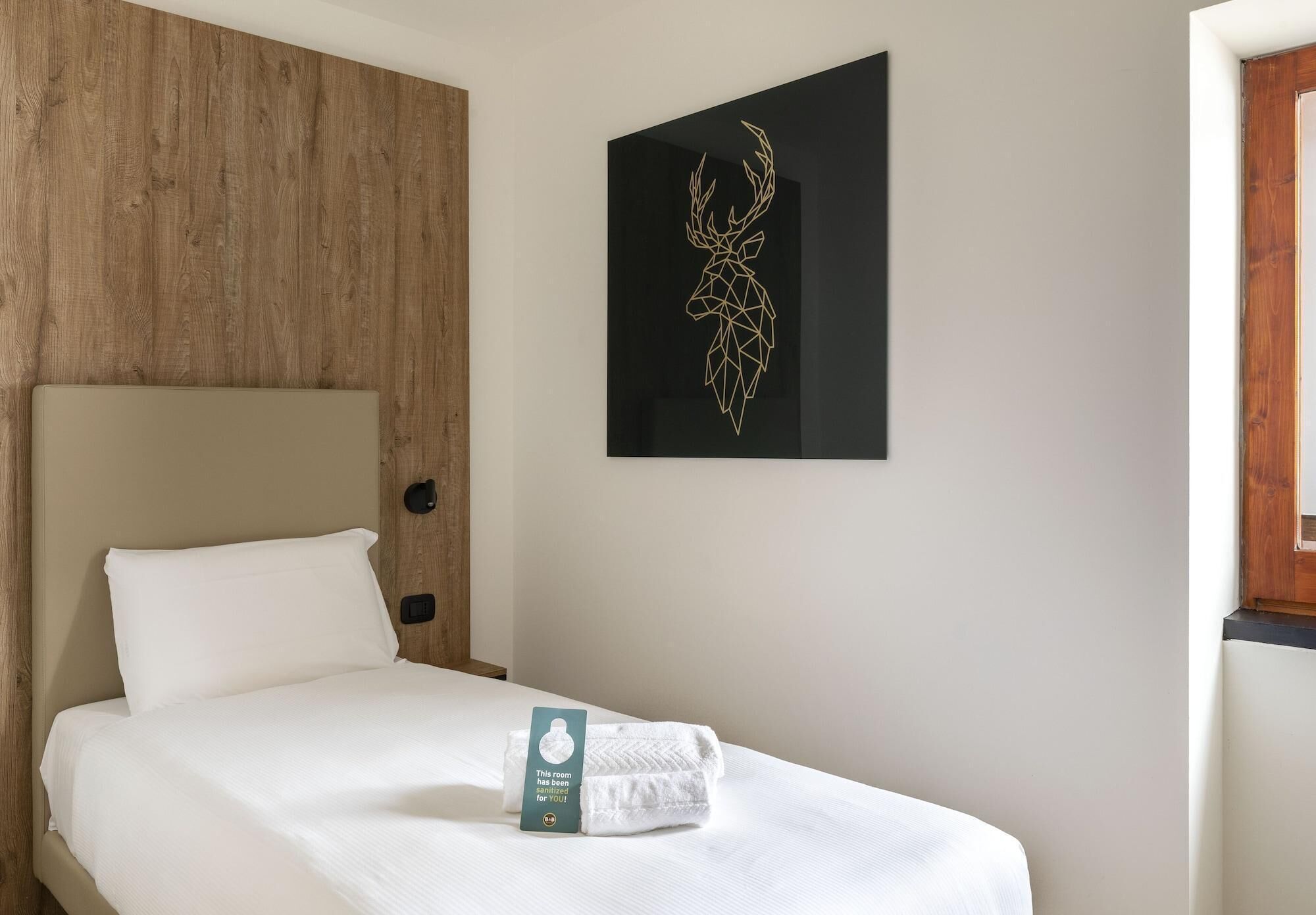 A neatly made bed with a folded towel and a tag, a wooden headboard, and a wall-mounted artwork of a deer head.