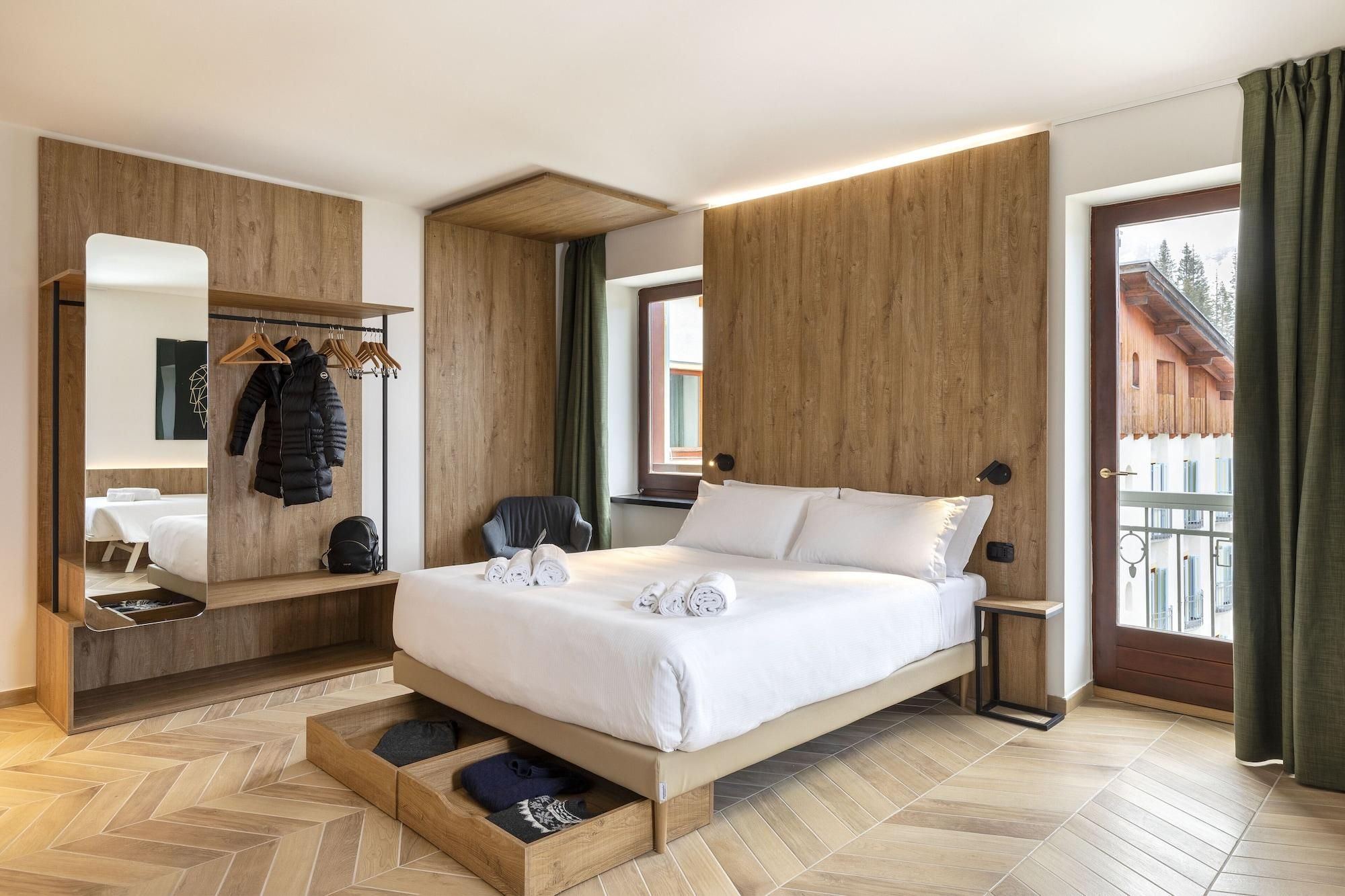 A modern hotel room with a wooden bed frame, a built-in wardrobe, a seating area, and a view of the outside.