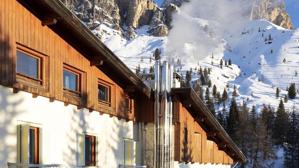 A wooden building with multiple windows and balconies, set against a backdrop of snow-covered mountains and clear blue sky.