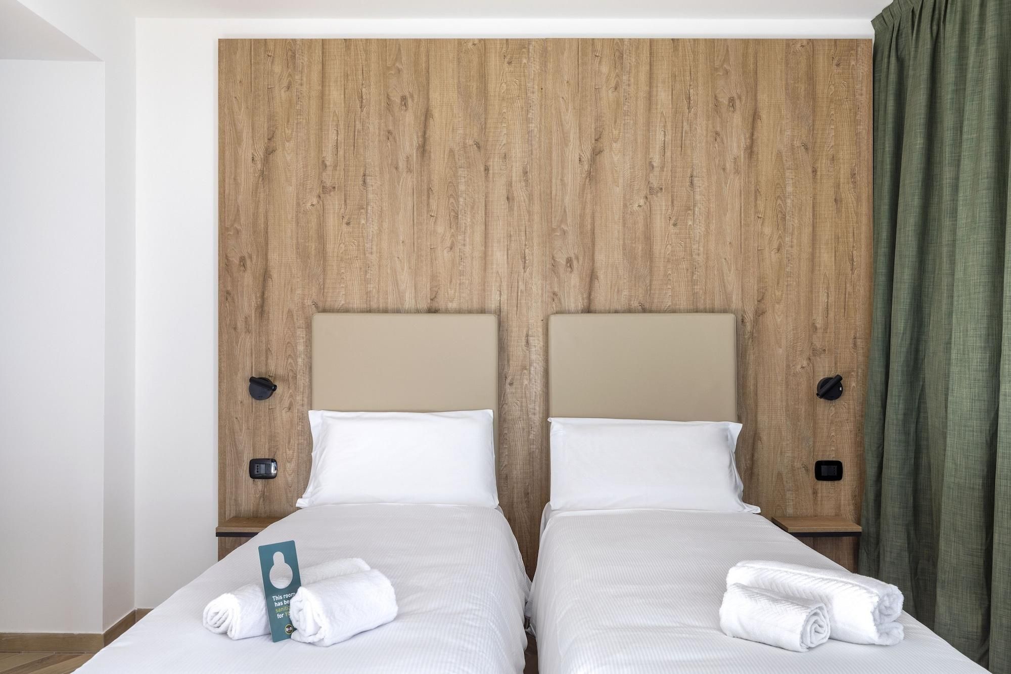 A double bed with two pillows and a folded towel, a wooden headboard, and a nightstand with a phone.