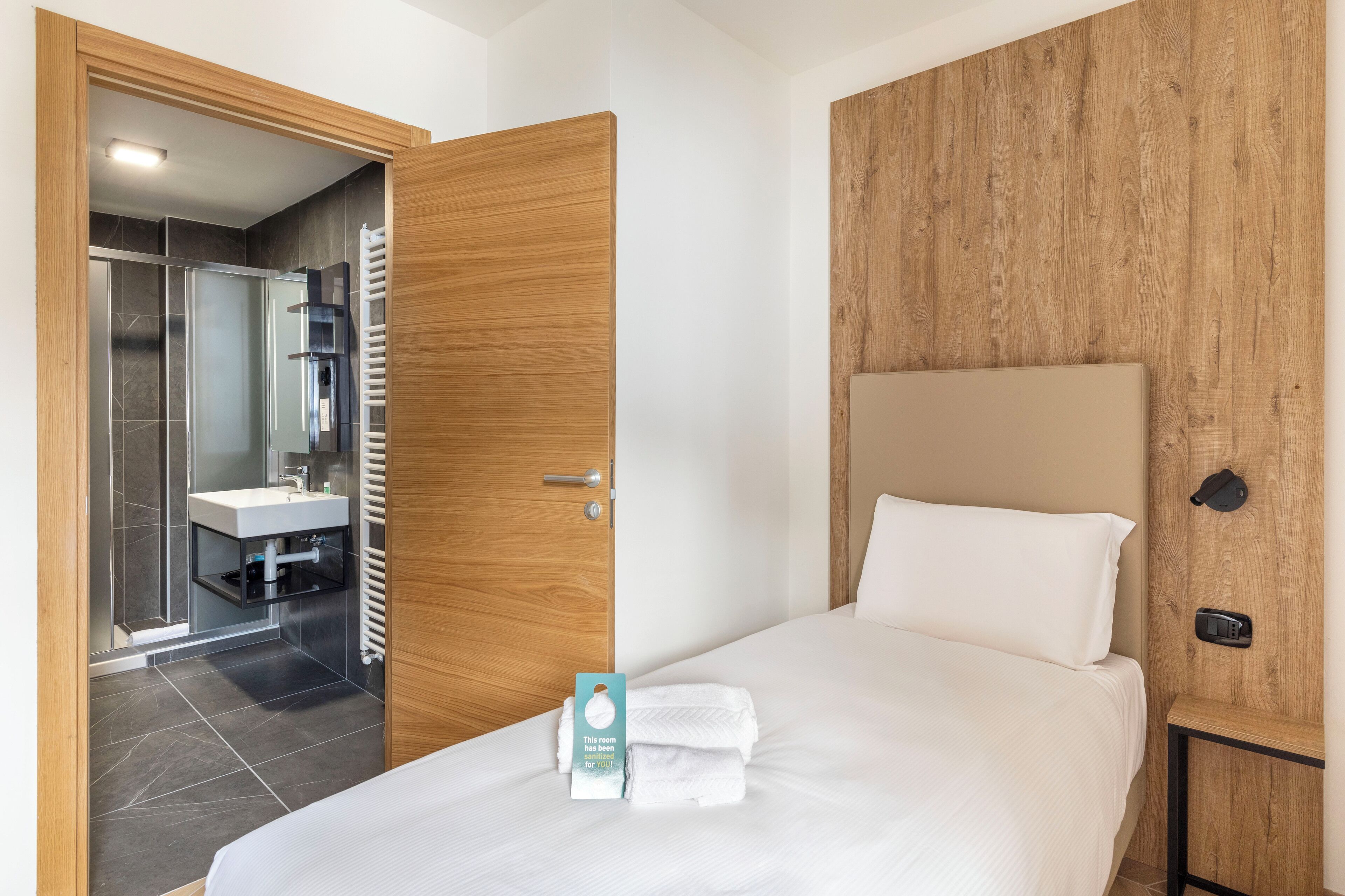 A hotel room with a wooden headboard, a bed with white linens, a bedside table, and a view into the bathroom.