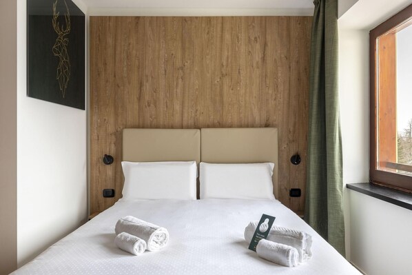 Habitación de hotel con cabecera de madera, dos almohadas blancas, cama con sábanas blancas y dos toallas enrolladas sobre la cama.