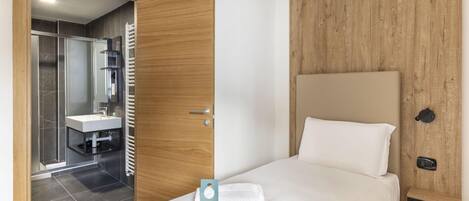 A hotel room with a bed, a wooden headboard, a bathroom with a shower, and a towel on the bed.