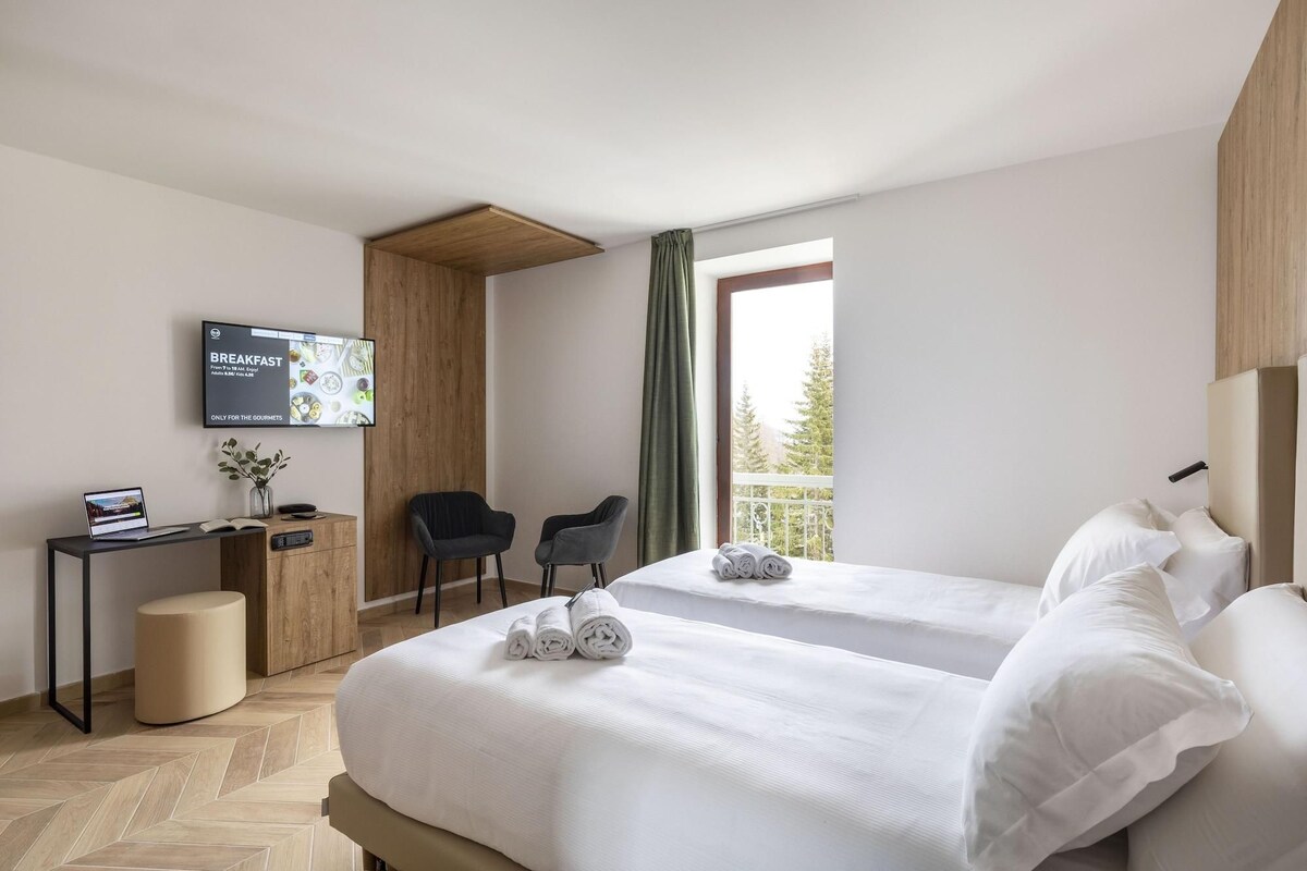 A hotel room with two beds, a desk with a laptop, a TV, and a view of trees through a window.