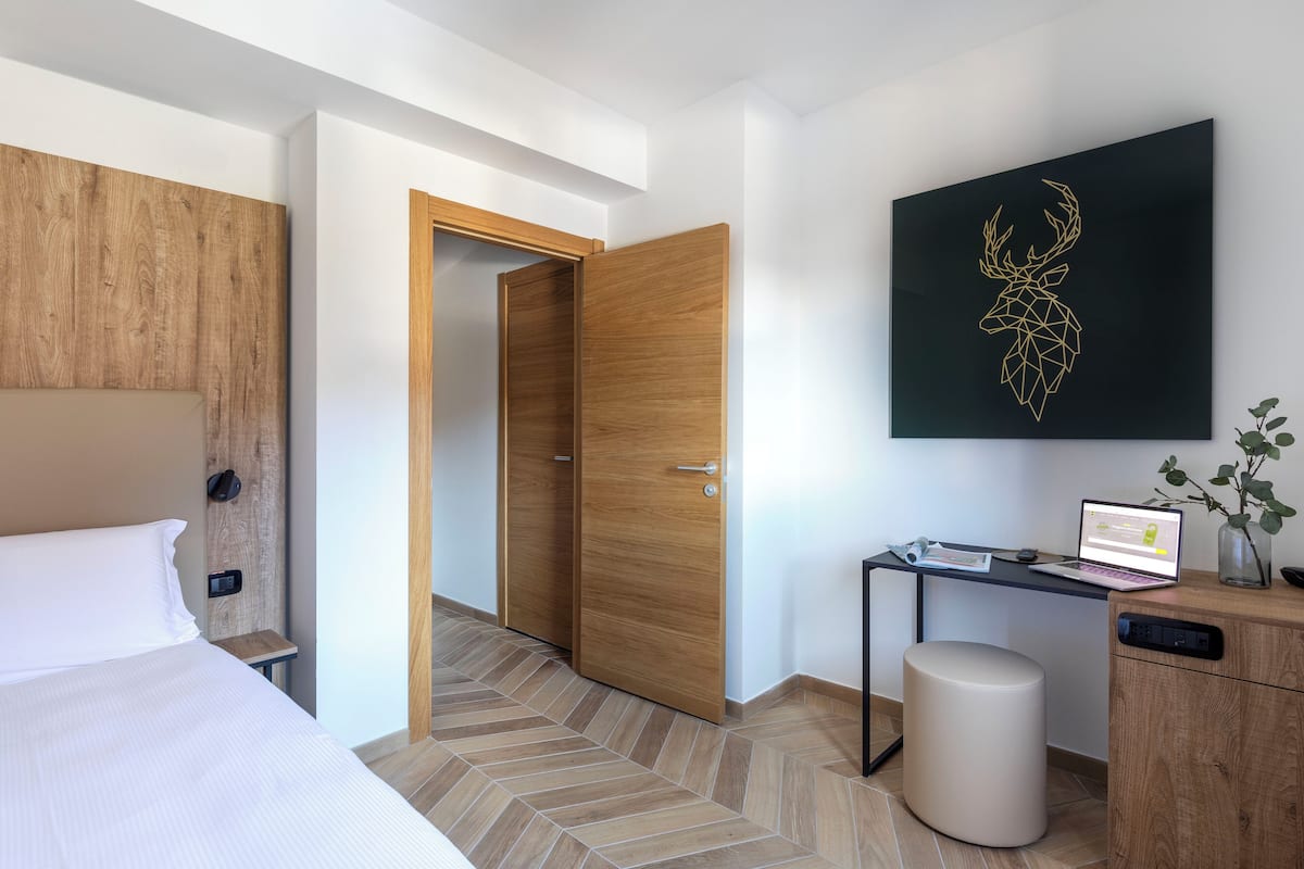 Ein modernes Hotelzimmer mit einem hölzernen Kopfteil, einem Bett mit weißer Bettwäsche, einem Schreibtisch mit einem Laptop, einer kleinen Topfpflanze und einem Wandbild eines Rehgehörns.
