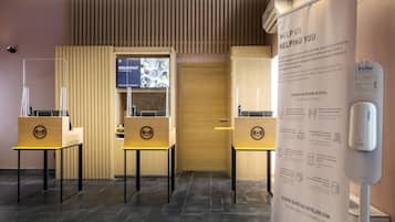 Uma área de recepção de hotel com um balcão de madeira, um painel de menu digital e um cartaz com orientações sobre COVID-19.