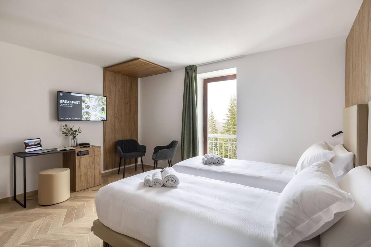 Ein Hotelzimmer mit zwei Betten, einem Schreibtisch mit einem Laptop, einem Fernseher, der Frühstücksangebote zeigt, und einem Fenster mit Blick auf Bäume.