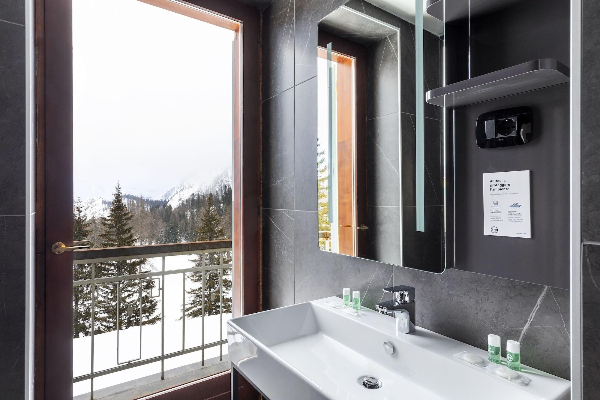 Ein modernes Badezimmer mit einem großen Spiegel, einem weißen Waschbecken und Blick auf eine verschneite Berglandschaft durch eine Balkontür.
