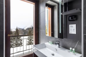 A modern bathroom with a large mirror, a white sink, and a view of a snowy mountain landscape through a balcony door. - B&B Hotel Passo Tre Croci Cortina (Cortina d'Ampezzo)