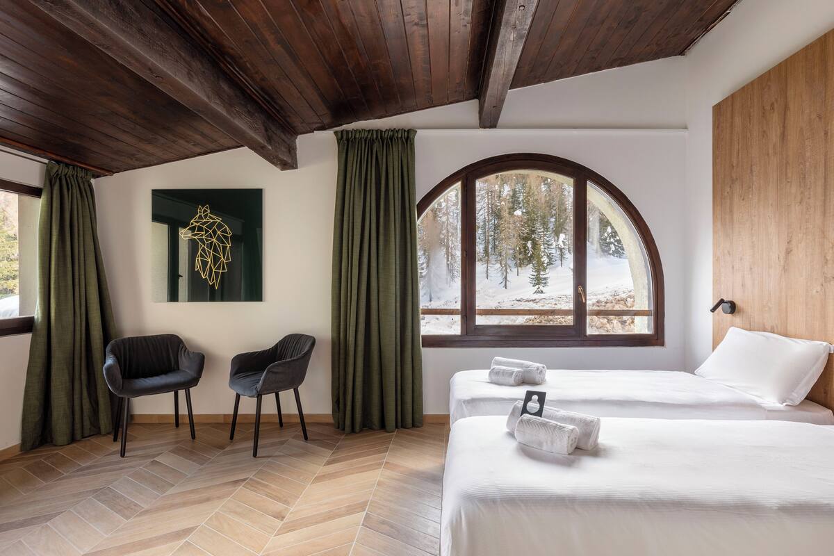 Ein Zimmer mit zwei Betten, einer Holzbalkendecke, einem großen Bogenfenster mit Blick auf Bäume und einem geometrischen Wandbild.