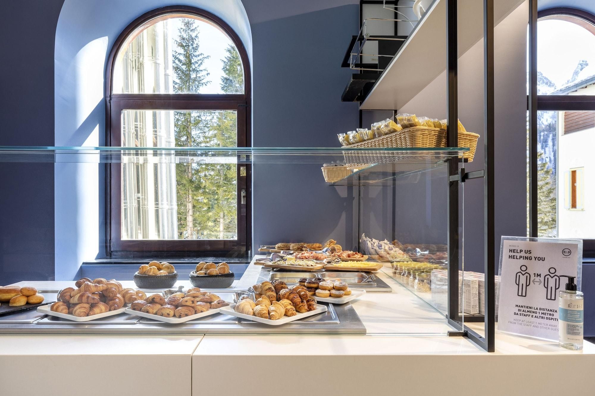Un buffet de petit-déjeuner proposant diverses pâtisseries et pains présentés dans une vitrine, avec un panneau indiquant les mesures de distanciation sociale.