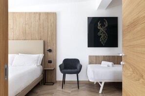 Una habitación de hotel moderna con una cama de madera, una mesita de noche pequeña, una silla negra y una cama blanca con cabecera de madera.