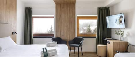 Habitación de hotel con cama, dos sillas, escritorio, televisor y ventana con vista a árboles.