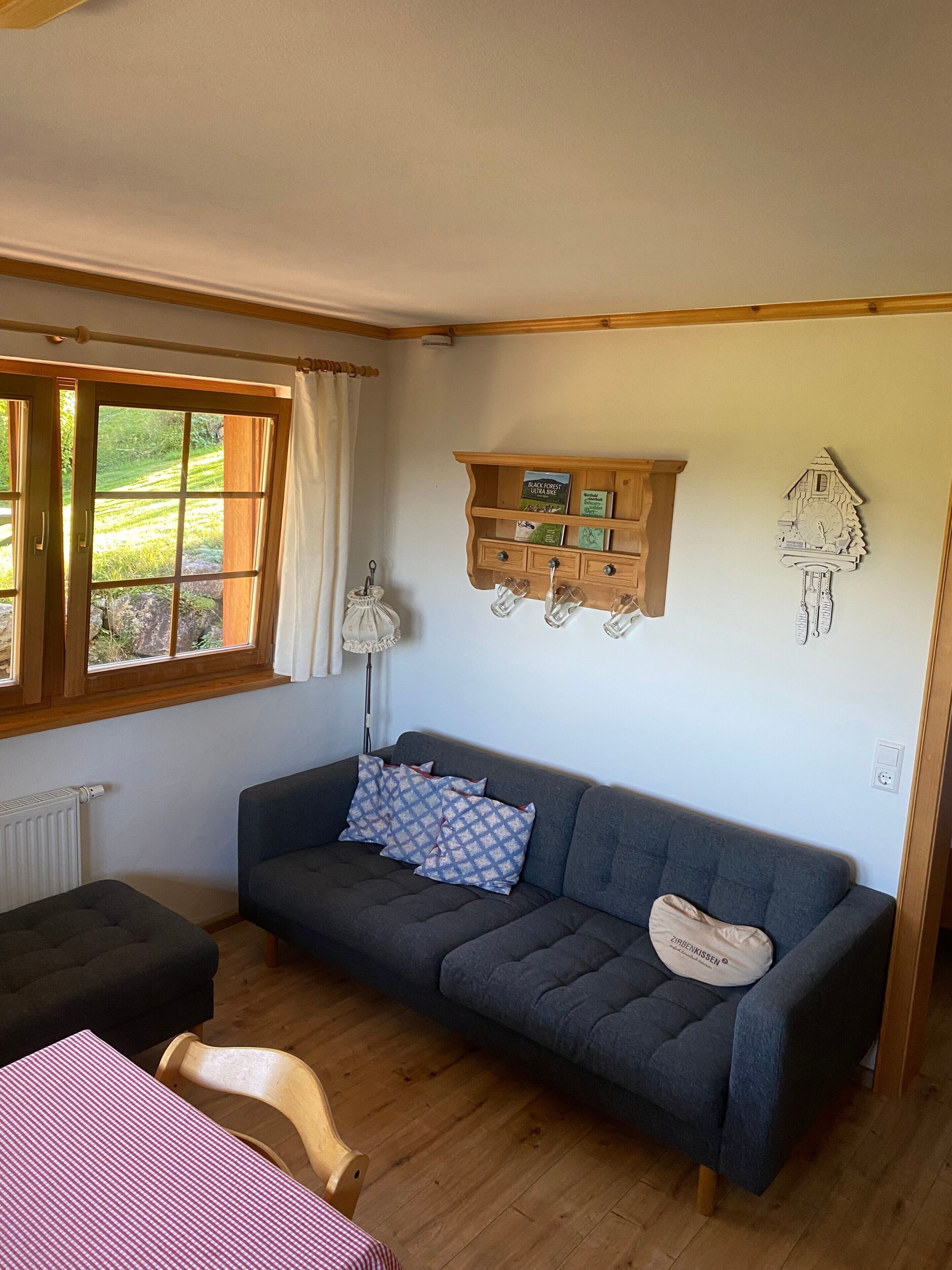 A room with a sofa, a table, a shelf with books, a clock, and a window with curtains.